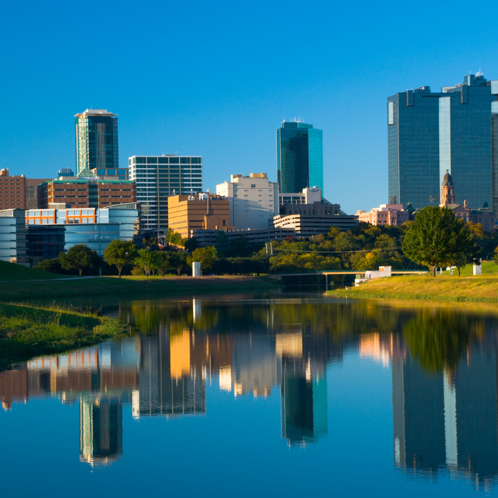 Fort Worth, Texas skyline with modern buildings and reflections in the Trinity River under a clear blue sky.