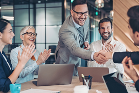 Smiling professionals in business attire celebrate a successful meeting in a modern office, with two shaking hands across the table as colleagues applaud. Image represents blog covering, what is an executive?
