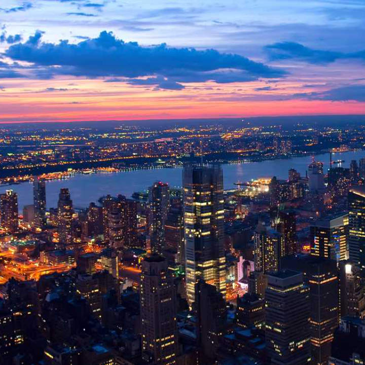 A high-rise cityscape at dusk with illuminated office buildings, a glowing sunset over the river, and city lights stretching into the horizon—representing the thriving professional environment served by Beacon Hill Parsippany’s temp NJ staffing solutions.