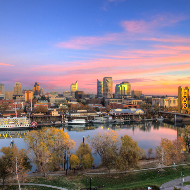 A colorful sunset view of downtown Sacramento, California, featuring modern office buildings, the waterfront, historic riverboats, and the Tower Bridge—highlighting the city served by temp staffing and consulting services.
